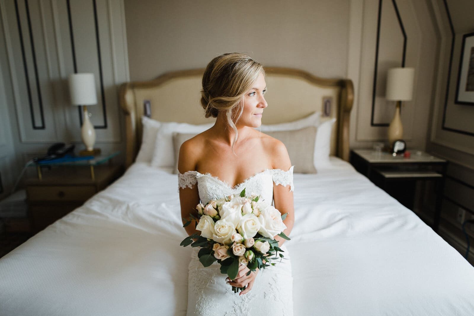 Une jeune mariée dans sa robe assise sur son lit fait de draps blancs et tenant dans ses mains un bouquet de fleurs roses pastels et blanches