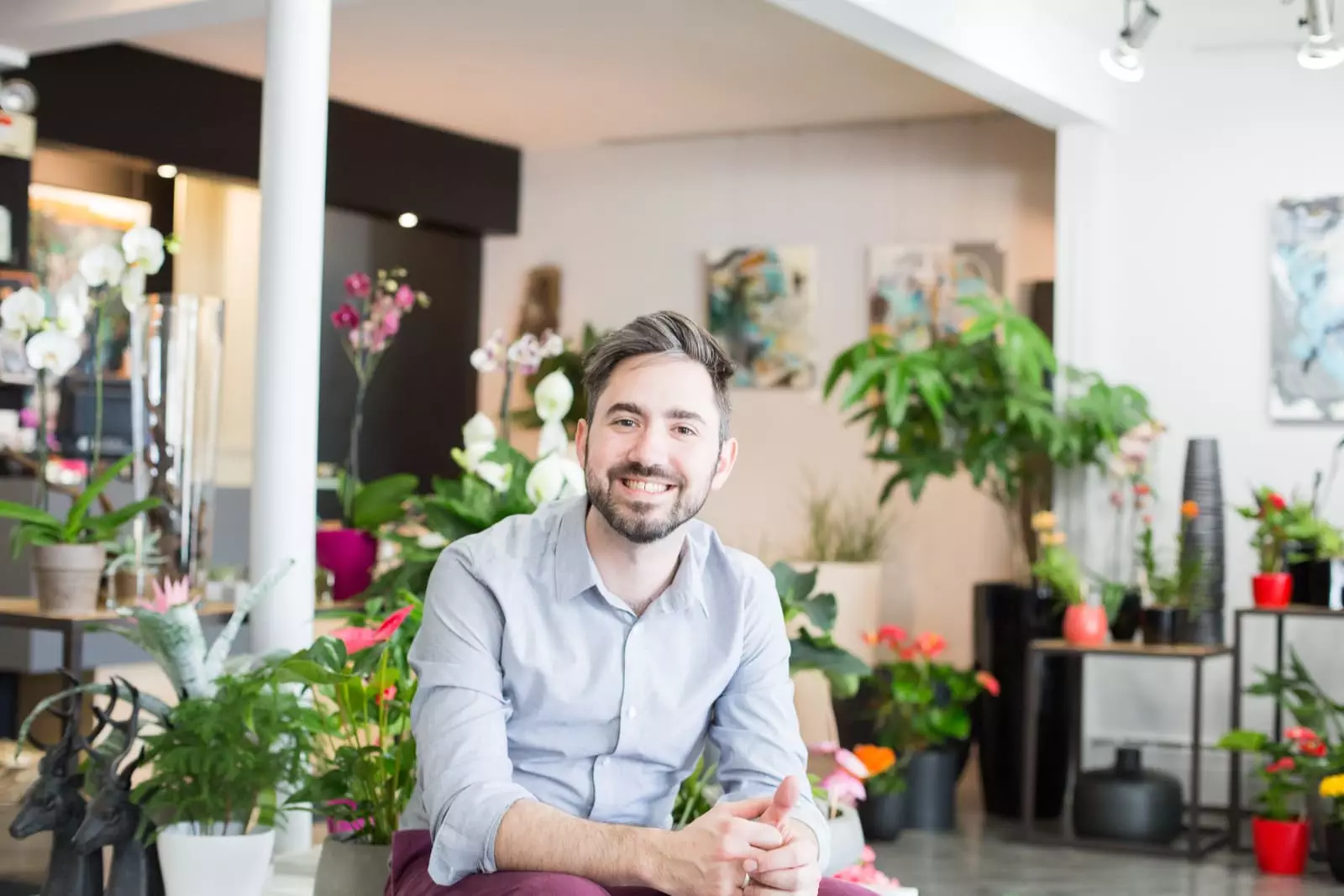 Homme qui souris au photographe assis parmi des fleurs et des plantes vertes